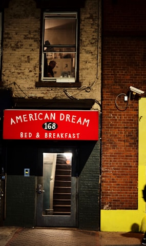 A building facade features a red sign with white lettering reading 'AMERICAN DREAM 168 BED & BREAKFAST' above a door with a glass panel revealing interior stairs. A window on the upper floor shows part of an industrial-looking room with a person visible, creating a scene suggestive of urban life. The walls are made of brick and painted surfaces, and there is a security camera mounted on the wall.