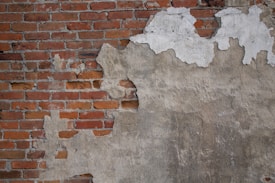 A wall with peeling plaster revealing red bricks beneath. The plaster is mostly worn away on the left side, exposing more of the textured brick surface. The remaining plaster is discolored and weathered, giving a rugged appearance.