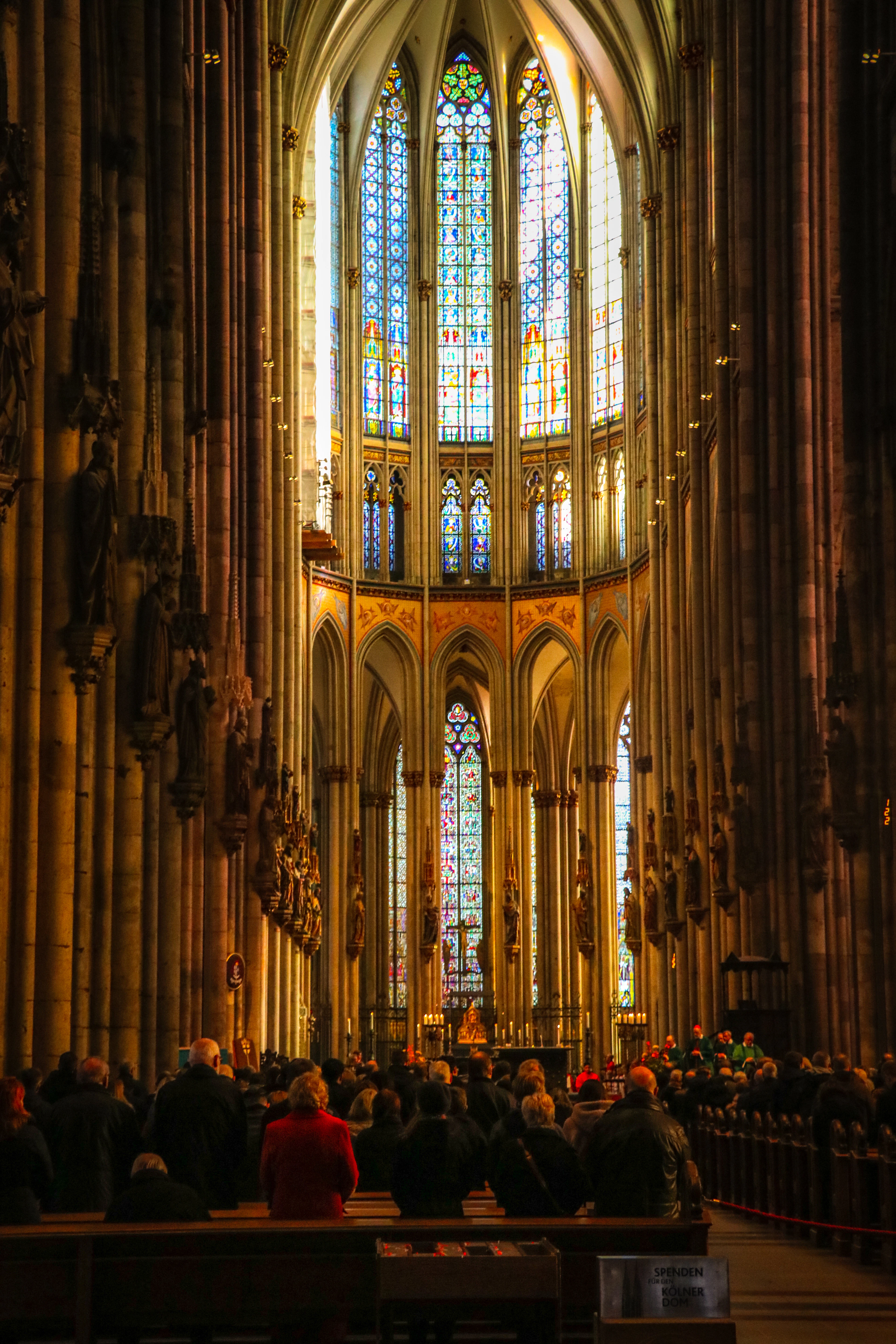 The soaring choir of Cologne Cathedral seen down the nave, its towering medieval stained glass glowing blue and gold above the lit high altar while a congregation stands in the shadowed pews below.