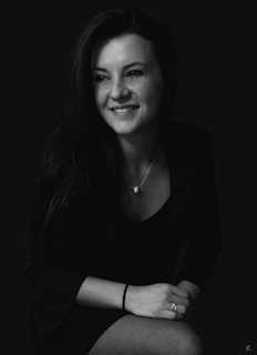 A black and white portrait of a smiling woman with long dark hair. She wears a dark outfit and a pendant necklace. Her hands are resting on her lap, with a ring on one finger and a hair tie around her wrist.