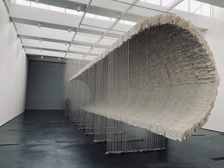 A large, sculptural installation hangs from the ceiling of a modern gallery space, composed of layered, textured material suspended by strings. The installation curves, creating a tunnel-like form that dominates the room. The gallery walls and floors are minimalistic and complement the artwork.