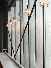 A row of industrial-style hanging light fixtures with metal cages emit a warm glow. These lights are suspended from a high ceiling along large vertical windows, which allow soft natural light to seep through. The space appears modern and minimalistic, with a long white countertop visible at the bottom left.