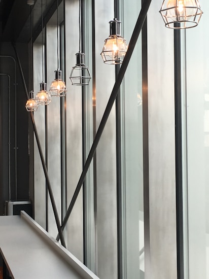 A row of industrial-style hanging light fixtures with metal cages emit a warm glow. These lights are suspended from a high ceiling along large vertical windows, which allow soft natural light to seep through. The space appears modern and minimalistic, with a long white countertop visible at the bottom left.