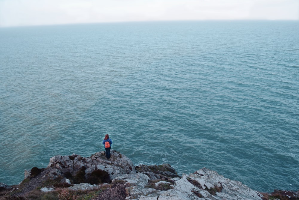 woman in blue jacket with pink backpack standing on cliff over the sea