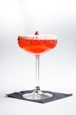 A vibrant cocktail in a coupe glass with a frothy top garnished with a berry, against a minimalist white background. The rim of the glass is adorned with red sugar or salt, and the drink sits on a black napkin.