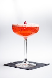 A vibrant cocktail in a coupe glass with a frothy top garnished with a berry, against a minimalist white background. The rim of the glass is adorned with red sugar or salt, and the drink sits on a black napkin.