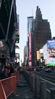 Tall buildings with bright billboards and advertisements line a busy urban street. Pedestrians and some vehicles are visible, with orange barriers partially blocking part of the sidewalk. The sky is clear, and the overall setting is a bustling city environment.