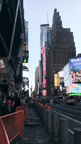 Tall buildings with bright billboards and advertisements line a busy urban street. Pedestrians and some vehicles are visible, with orange barriers partially blocking part of the sidewalk. The sky is clear, and the overall setting is a bustling city environment.
