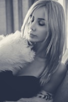 A woman with long hair is resting her head on a fluffy, light-colored pillow or fur, her eyes closed and face calm. She wears a strapless black top, and the setting is softly lit, contributing to an intimate and serene atmosphere.