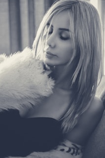 A woman with long hair is resting her head on a fluffy, light-colored pillow or fur, her eyes closed and face calm. She wears a strapless black top, and the setting is softly lit, contributing to an intimate and serene atmosphere.
