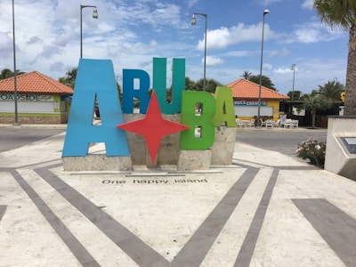 Large colorful sign spelling out 'ARUBA' with a red star in the middle, situated in a public area with paved surroundings and lampposts. Palm trees and buildings with red-tiled roofs are visible in the background, under a partly cloudy sky.