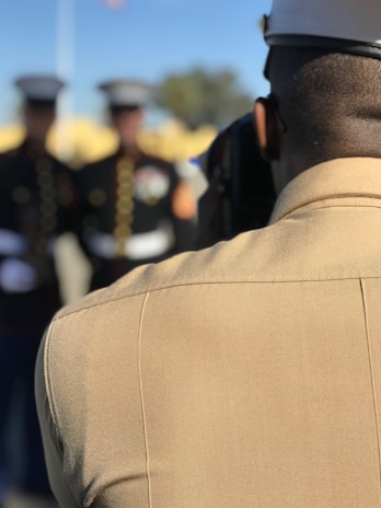 A uniformed individual stands in the foreground, wearing a cap and a khaki-colored outfit. Blurred figures in similar military attire are visible in the background, suggesting a formal or ceremonial setting.