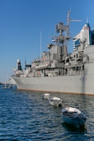 A large naval ship is docked in a harbor with several small white boats tethered alongside in the water. The ship is gray and equipped with various antennas and communication equipment. In the background, there are modern buildings, one of which has 'Hotel' written on it. The sky is clear with a deep blue hue.