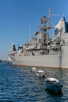 A large naval ship is docked in a harbor with several small white boats tethered alongside in the water. The ship is gray and equipped with various antennas and communication equipment. In the background, there are modern buildings, one of which has 'Hotel' written on it. The sky is clear with a deep blue hue.