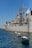 A large naval ship is docked in a harbor with several small white boats tethered alongside in the water. The ship is gray and equipped with various antennas and communication equipment. In the background, there are modern buildings, one of which has 'Hotel' written on it. The sky is clear with a deep blue hue.