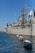 A large naval ship is docked in a harbor with several small white boats tethered alongside in the water. The ship is gray and equipped with various antennas and communication equipment. In the background, there are modern buildings, one of which has 'Hotel' written on it. The sky is clear with a deep blue hue.
