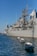 A large naval ship is docked in a harbor with several small white boats tethered alongside in the water. The ship is gray and equipped with various antennas and communication equipment. In the background, there are modern buildings, one of which has 'Hotel' written on it. The sky is clear with a deep blue hue.