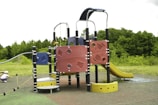 A colorful playground structure featuring various climbing elements, including panels with geometric patterns, a slide, and a climbing rope. The structure is set in an open area surrounded by greenery, with a child playing near one of the features.