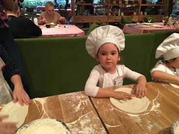 Children wearing white chef hats are preparing dough at a wooden table covered in flour. The background features a restaurant setting with people dining at tables with checkered tablecloths.
