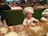 Children wearing white chef hats are preparing dough at a wooden table covered in flour. The background features a restaurant setting with people dining at tables with checkered tablecloths.