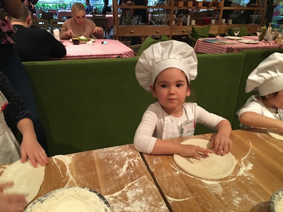 Children wearing white chef hats are preparing dough at a wooden table covered in flour. The background features a restaurant setting with people dining at tables with checkered tablecloths.