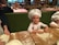 Children wearing white chef hats are preparing dough at a wooden table covered in flour. The background features a restaurant setting with people dining at tables with checkered tablecloths.