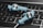 Two blue in-ear headphones with cables rest on a laptop keyboard. The keyboard keys are backlit in white, and the headphones have a sleek design with a prominent logo on the earpiece.