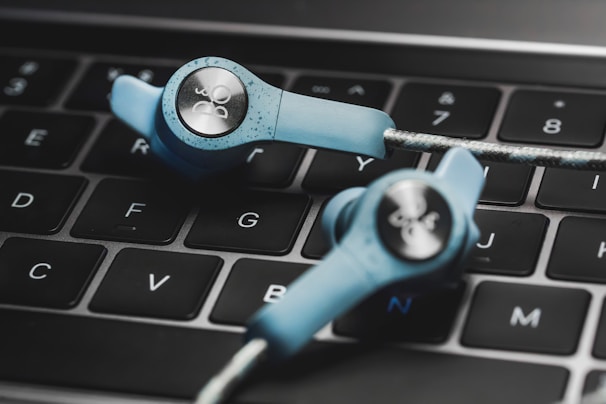 Two blue in-ear headphones with cables rest on a laptop keyboard. The keyboard keys are backlit in white, and the headphones have a sleek design with a prominent logo on the earpiece.