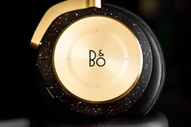 A close-up view of a stylish headphone earcup featuring a sleek gold and black design with a circular logo in the center. The earcup has speckled gold accents on a dark background, emphasizing a luxurious and modern aesthetic.