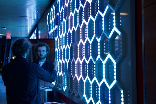 A person is holding a mirror, which reflects their image, in front of a brightly illuminated wall with a geometric hexagonal pattern made of blue and white lights. The environment appears modern with a polished ceiling and walls.