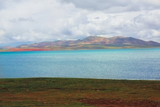 A tranquil lake with clear blue water is surrounded by expansive green fields. In the background, a range of softly colored mountains rises under a cloudy sky.