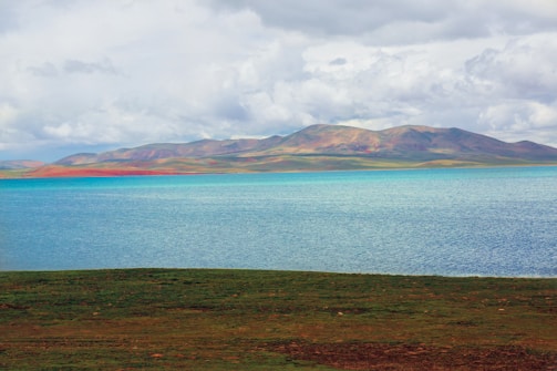 A tranquil lake with clear blue water is surrounded by expansive green fields. In the background, a range of softly colored mountains rises under a cloudy sky.