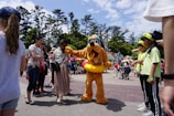 A theme park scene where a person in a Pluto costume, complete with a yellow float around the waist, interacts with a group of visitors. The background includes colorful buildings, tall trees, and a partly cloudy sky. People are casually dressed and appear to be enjoying the day.