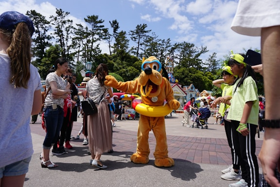 A theme park scene where a person in a Pluto costume, complete with a yellow float around the waist, interacts with a group of visitors. The background includes colorful buildings, tall trees, and a partly cloudy sky. People are casually dressed and appear to be enjoying the day.