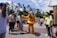 A theme park scene where a person in a Pluto costume, complete with a yellow float around the waist, interacts with a group of visitors. The background includes colorful buildings, tall trees, and a partly cloudy sky. People are casually dressed and appear to be enjoying the day.
