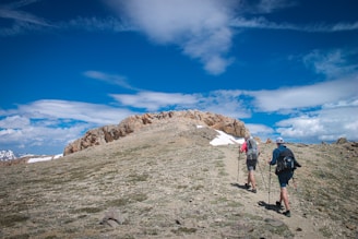 Two hikers with backpacks and trekking poles are walking up a rocky trail towards a summit. The surrounding landscape is a mix of sparse grass and rocky terrain, with patches of snow visible. The sky above is a bright blue with scattered clouds, giving a sense of open space and high altitude.