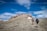 Two hikers with backpacks and trekking poles are walking up a rocky trail towards a summit. The surrounding landscape is a mix of sparse grass and rocky terrain, with patches of snow visible. The sky above is a bright blue with scattered clouds, giving a sense of open space and high altitude.