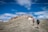 Two hikers with backpacks and trekking poles are walking up a rocky trail towards a summit. The surrounding landscape is a mix of sparse grass and rocky terrain, with patches of snow visible. The sky above is a bright blue with scattered clouds, giving a sense of open space and high altitude.