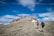 Two hikers with backpacks and trekking poles are walking up a rocky trail towards a summit. The surrounding landscape is a mix of sparse grass and rocky terrain, with patches of snow visible. The sky above is a bright blue with scattered clouds, giving a sense of open space and high altitude.