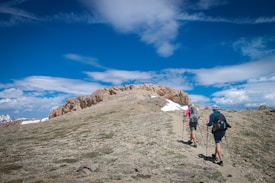 Two hikers with backpacks and trekking poles are walking up a rocky trail towards a summit. The surrounding landscape is a mix of sparse grass and rocky terrain, with patches of snow visible. The sky above is a bright blue with scattered clouds, giving a sense of open space and high altitude.