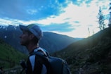 A person wearing outdoor gear, including a backpack and cap, appears to be hiking in a mountainous area during either sunrise or sunset. The sky is mostly cloudy with some visible patches of blue, and the mountains are covered in evergreen trees.
