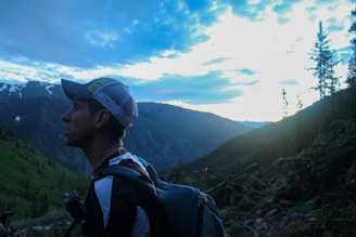 A person wearing outdoor gear, including a backpack and cap, appears to be hiking in a mountainous area during either sunrise or sunset. The sky is mostly cloudy with some visible patches of blue, and the mountains are covered in evergreen trees.