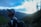 A person wearing outdoor gear, including a backpack and cap, appears to be hiking in a mountainous area during either sunrise or sunset. The sky is mostly cloudy with some visible patches of blue, and the mountains are covered in evergreen trees.