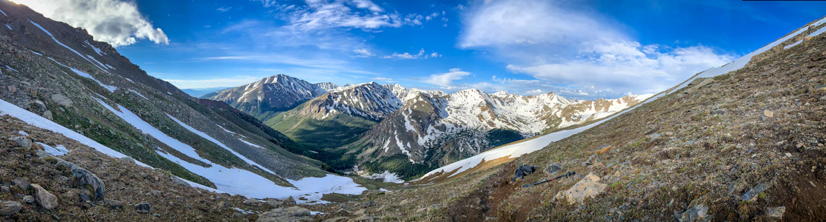 Snow-streaked peaks above a green valley, seen from a high, rocky slope on the Mount Massive Trail near Leadville