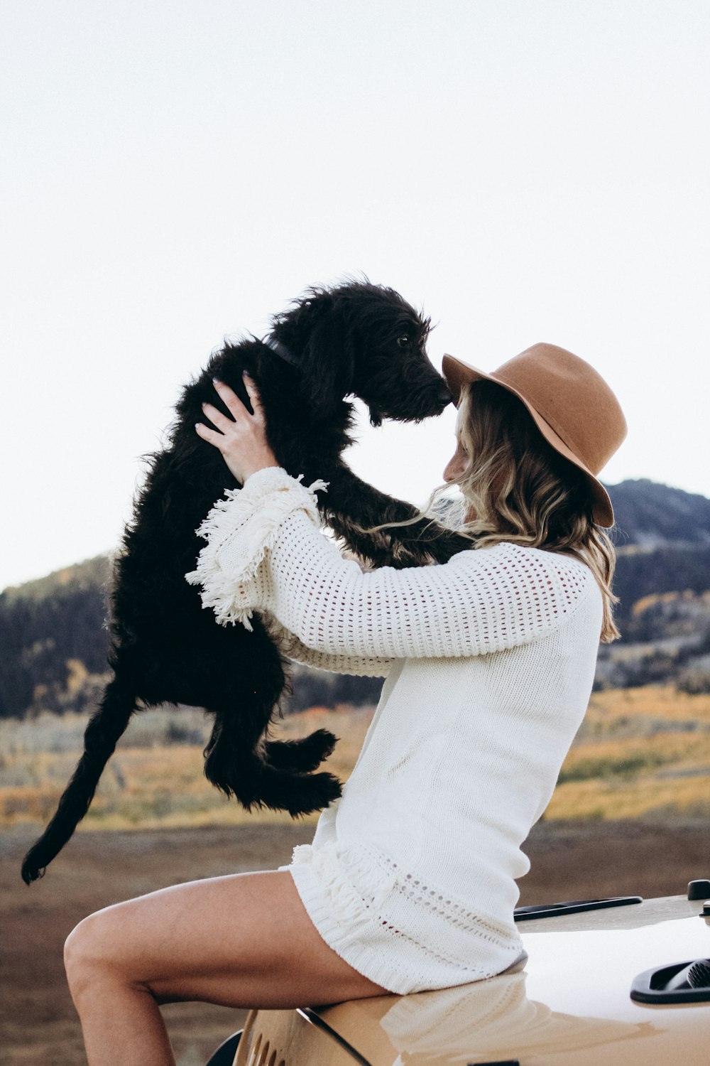 woman in white sweater dress sitting on hood of parked beige Jeep Wrangler holding up balck dog