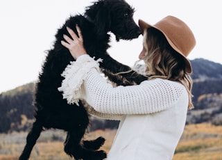 woman in white sweater dress sitting on hood of parked beige Jeep Wrangler holding up balck dog