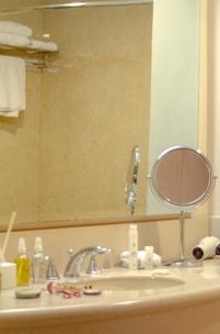 A bathroom countertop with various toiletry items arranged around a sink. A round magnifying mirror stands next to a wall-mounted hairdryer. Several bottles and a toothbrush container are placed around the faucet. On the left, folded white towels are neatly arranged on a wall shelf against a beige tiled wall.
