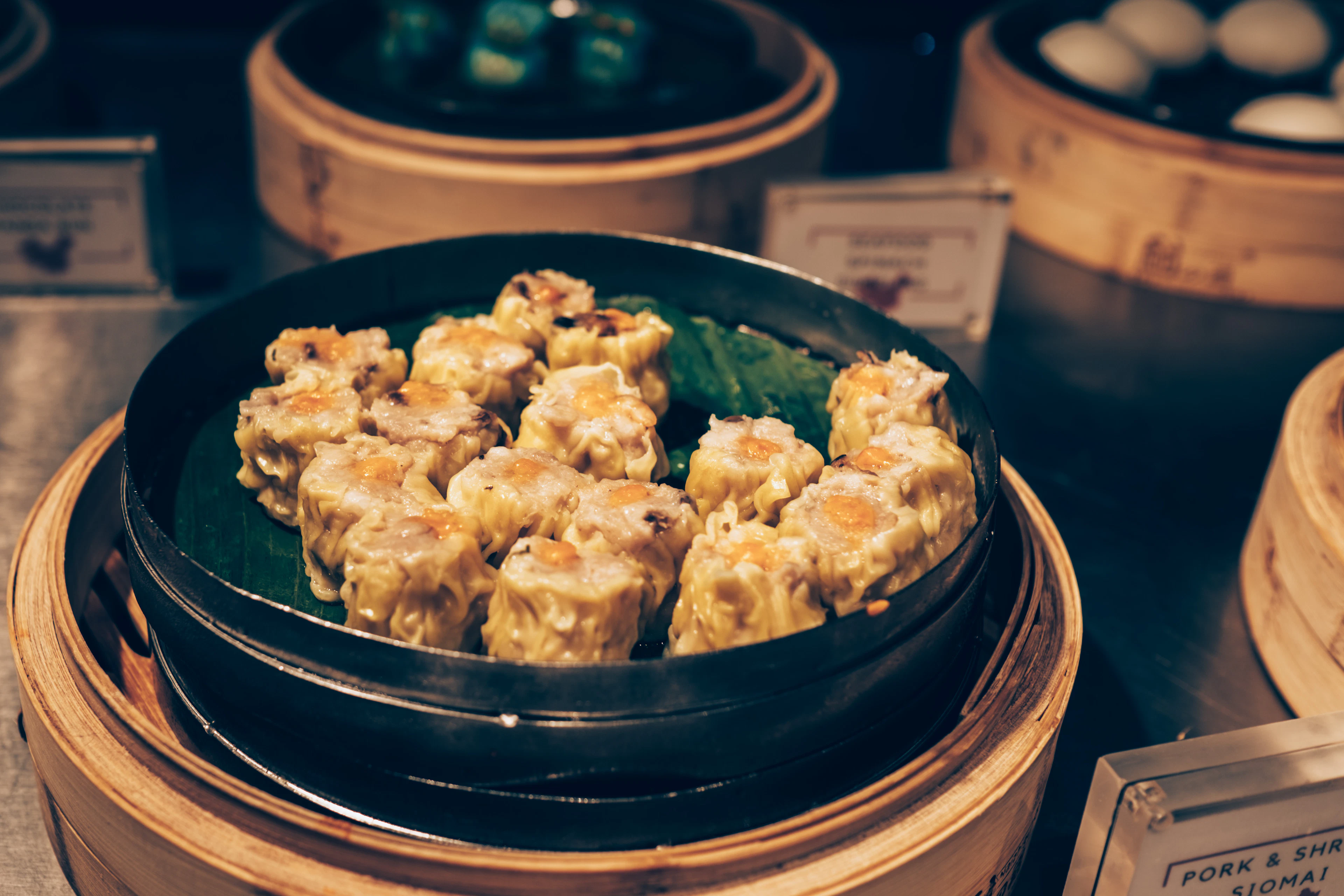 Siu mai pork dumplings on a leaf-lined plate set inside stacked bamboo steamers at a dim sum counter.