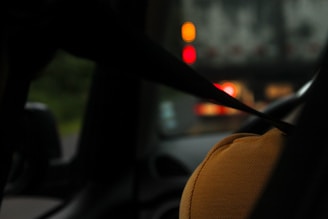 A person wearing a yellow shirt is sitting in a car with a seat belt fastened. The focus is on the shoulder and seat belt, with blurred traffic lights visible through the windshield.