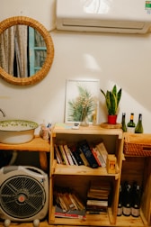 A cozy interior corner with a circular wicker-framed mirror reflecting a window with curtains. Below the mirror, a wooden shelf holds a variety of books and a framed botanical print. A potted plant on the shelf adds greenery. Above, an air conditioning unit is mounted on the wall. To the side, bottles of wine are neatly arranged, and a small fan sits near a sink with a decorative bowl.